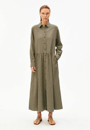 Femme portant une robe longue à manches longues, à carreaux verts et marron, boutonnée sur le devant, debout avec une main dans la poche.