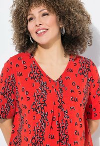 Blusa corta roja de manga corta con escote en V, que presenta un patrón texturizado de letras negras en diversas orientaciones.