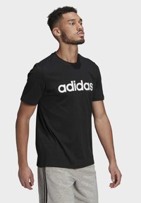 Mustat puuvillat-paita, jossa on suuri valkoinen "adidas"-logo etuosassa, pyöreä kaula-aukko ja lyhyet hihat, yhdistettynä harmaisiin collegehousuihin.