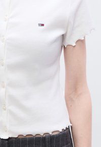 Λευκή κοντομάνικη ζακέτα με ριμπ ύφανση και κυματιστό τελείωμα, με μικρό κεντημένο λογότυπο στο αριστερό στήθος. Υφή υφάσματος.