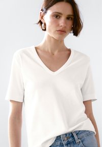 Biała koszulka typu V-neck z gładkiego materiału, z krótkimi rękawami. Noszona z jasnoniebieskimi dżinsami. Casualowy krój, minimalistyczny wzór, brak widocznych motywów.