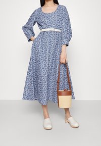 Blau-weiße Paisley-muster lange Kleid mit Rundhalsausschnitt und Puffärmeln, kombiniert mit transparenten Mules und einer braunen Stroh-Handtasche.