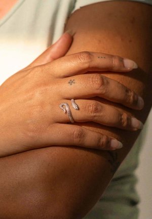 Silberner Schlangenring mit strukturiertem Design, getragen an einer Hand mit hellen, neutralen Hautton, zusammen mit mehreren einfachen Tattoos.