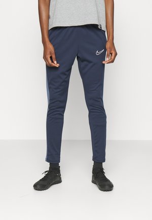 Persoon draagt donkerblauwe Nike trainingsbroek met wit logo, zwarte sneakers en grijs shirt, staand tegen een witte achtergrond.