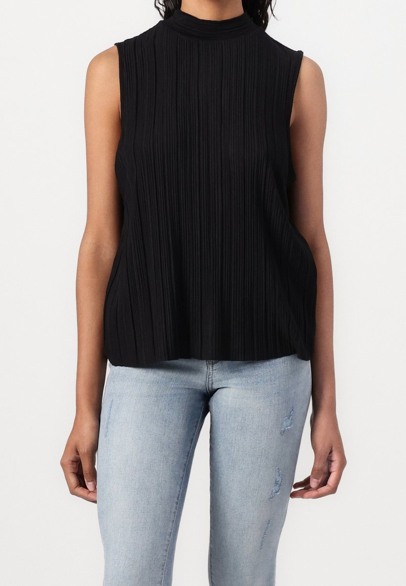 Kobieta w bezrękawnikowej, czarnej, plisowanej bluzce oraz jasnoniebieskich, podartych, dopasowanych dżinsach, stojąca na jednolitym tle.