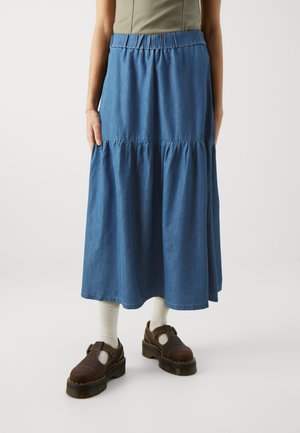 Denim svārki ar elastīgu jostasvietu, pakāpenisku dizainu un plūstošu siluetu. Uzvilkti ar brūniem apaviem un gaišām zeķēm.