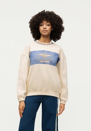 Νέα γυναίκα με σγουρά μαλλιά που φοράει μπεζ φούτερ με μπλε λογότυπο Billabong και σκούρο μπλε παντελόνι με λευκές ρίγες, στέκεται μπροστά σε απλό φόντο.