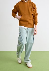 Persona vestida con una sudadera marrón, jeans cargo azul claro y zapatillas blancas, caminando sobre un suelo verde con un fondo de pared blanca.