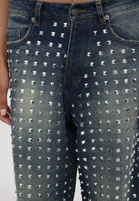 Un pantalon en denim orné de nombreux petits clous en forme de pyramide argentés couvrant la partie avant de la cuisse, porté par une personne avec la main reposant à côté de la jambe.