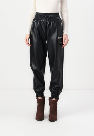 Nahaufnahme einer Person, die schwarze Jogger aus Kunstleder mit elastischen Bündchen und Reißverschlusstaschen trägt, kombiniert mit braunen Wildleder-Stiefeletten.