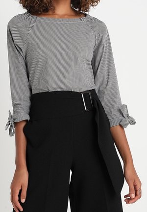 Femme portant un haut rayé vertical noir et blanc avec des poignets noués aux manches et un pantalon noir taille haute avec une ceinture large et une boucle.