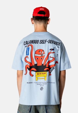 Person iført lyseblå T-shirt med rød blæksprutte, der holder køkkenredskaber, og tekst "Calamari Self-Service" og "Mad er folks første prioritet."