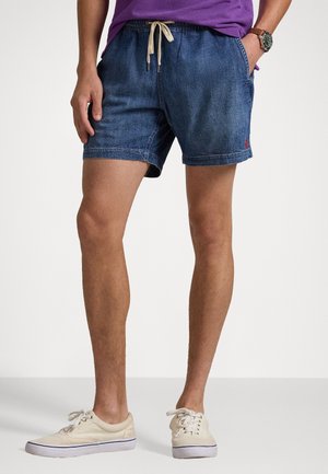 Man draagt blauwe denimshorts met trekkoord, paars shirt, beige sneakers en polshorloge, staand tegen een witte achtergrond.