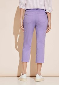 Liliowe spodnie o kroju capri wykonane z lekkiego materiału, z dwoma tylnymi kieszeniami i obszytymi mankietami, zestawione z białymi trampkami.