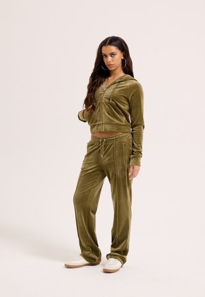 Žena s dlhými tmavými vlasmi v olivovo zelenej zamatovej mikine na zips a zodpovedajúcich nohaviciach, s bielymi teniskami, stojaca pred jednofarebným pozadím.