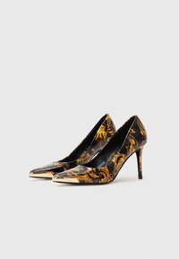 Černé lakované lodičky na vysokém podpatku se zlatým květinovým vzorem, špičatou špičkou a kovovým akcentem na špičce. Úzký design stiletto podpatku.