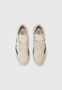 Beige mesh sneakers with a textured finish, green and black accents, and cream laces; includes a logo label on the tongue.