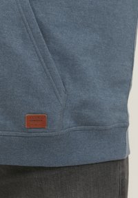 Sudadera de algodón azul con dobladillo y puños acanalados, con una etiqueta de cuero marrón cosida en la parte inferior izquierda. Diseño texturizado y casual.
