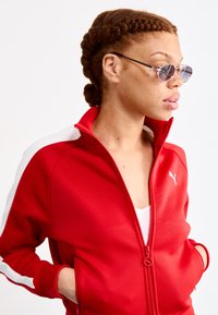 Crvena zip-up trenerka jakna s bijelim trakama sa strane, s visokim ovratnikom i teksturiranim materijalom. Okršena malim bijelim logotipom na prsima.