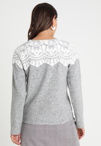 Femme aux cheveux foncés portant un pull gris avec une dentelle florale blanche sur le haut du dos et une jupe à carreaux, debout de dos.