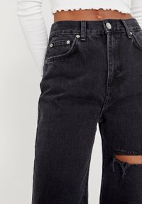 Jean en denim noir taille haute, avec une déchirure usée au niveau du genou, ferrures argentées et design classique à cinq poches.