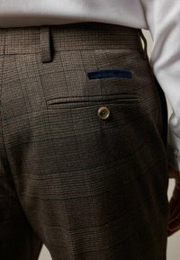 Pantaloni brown în carouri cu o textură netedă, având un singur buzunar posterior, închidere cu nasture și un detaliu din material albastru deasupra buzunarului.