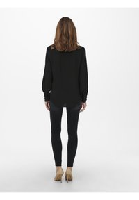 Schwarzes Langarmshirt mit lockerer Passform, strukturiertem Stoff und geknöpften Manschetten, kombiniert mit schwarzen Skinny-Jeans und beigen Absätzen.