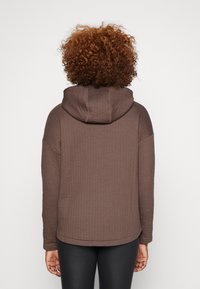 Sudadera con capucha de textura marrón y ajuste holgado, con una costura en la espalda y detalles acanalados. Se lleva sobre leggings negros.