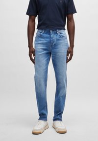 Man som bär ljusblå slim-fit jeans, vita sneakers och en marinblå t-shirt, stående mot en enfärgad grå bakgrund.