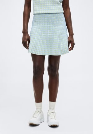 Partea inferioară a corpului unei persoane îmbrăcată cu o fustă plisată în carouri albastru deschis și galben, șosete albe și pantofi sport albi, pe un fundal simplu.