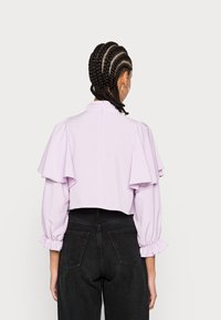 Blusa cropped lilás com mangas em camadas e babados, gola alta e fecho de zíper nas costas. Usada com jeans pretos de cintura alta.