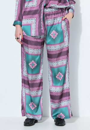Lockere Seidenhose mit lila, türkis und pinken geometrischen Mustern, getragen mit schwarzen Schuhen, Hände in den Taschen und Ringe zeigend.