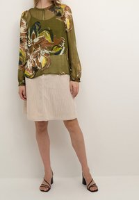 Blusa trasparente verde oliva con motivo floreale, abbinata a una gonna in cotone a coste color crema. Tacchi neri con cinturini completano l'outfit.