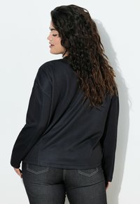 Schwarzes langärmliges Oberteil mit lockerem Schnitt und Rundhalsausschnitt. Weiche Textur, sichtbare Nahtakzente, kombiniert mit dunklen Jeans.