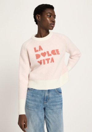 Young person wearing light pink sweater with "LA DOLCE VITA" in red letters and blue jeans, posing with one hand on hip.