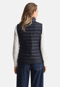 Tamsiai mėlyna pūkinė liemenė su vertikaliu siuvimu, aukštu apykakliu ir elastingu apačios kraštu. Dėvima virš baltos ilgomis rankovėmis marškinių, pasižyminčių laisvu siluetu.