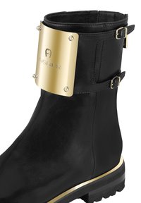 Mustat nahka nilkkurit, joissa on kultainen metallilevy, jossa on kaiverruslogo, koristeelliset soljet ja teksturoitu kumipohja kultaisella yksityiskohdalla.