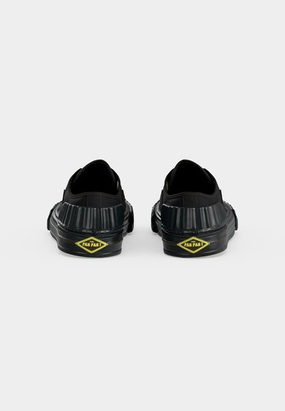 Paire de chaussures noires sans lacets avec talons rainurés et logo jaune en forme de losange portant l'inscription "THE FAN FAN !" sur un fond blanc uni.