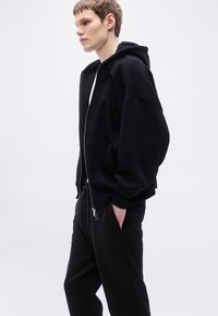 Μαύρο φούτερ με φερμουάρ, με απαλή υφή, φαρδιά γραμμή και τσέπες καγκουρό. Συνδυάζεται με μαύρο παντελόνι φόρμας, και τα δύο με μινιμαλιστικό σχεδιασμό.