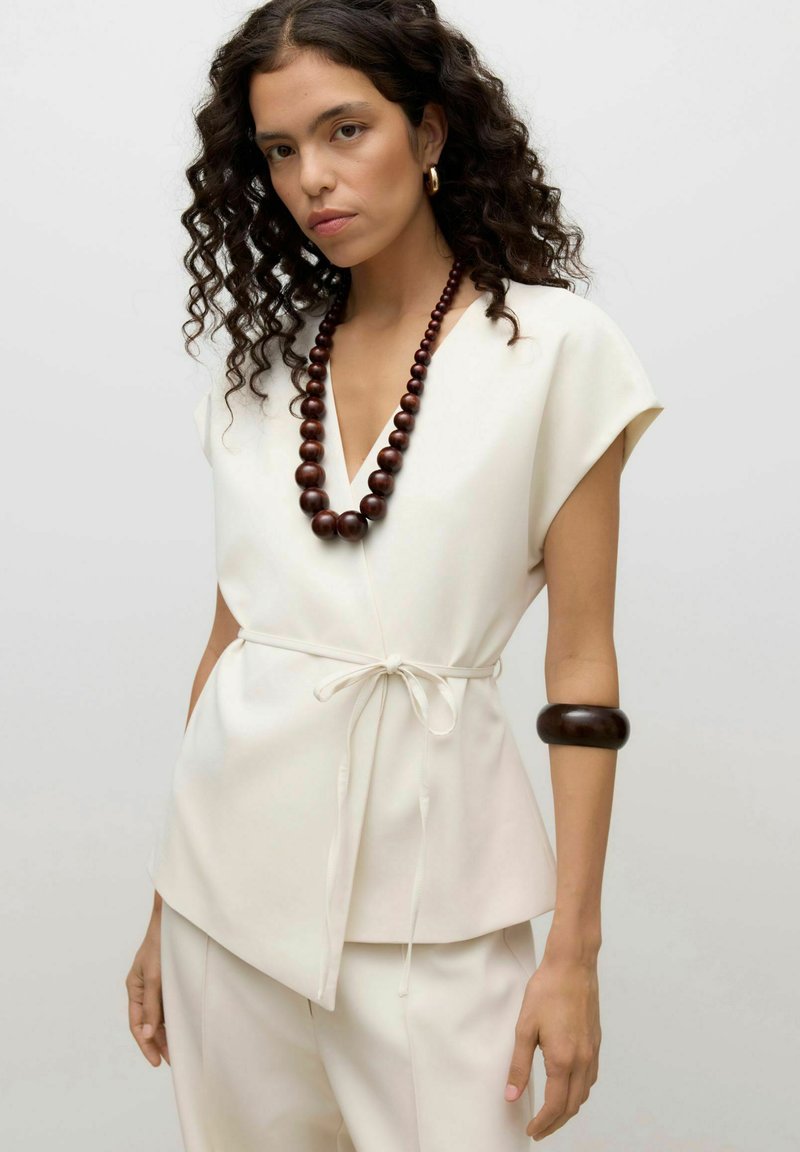 Femme aux cheveux bouclés portant un haut portefeuille sans manches écru et un pantalon, accessoirisée avec un grand collier et un bracelet en perles de bois foncé.