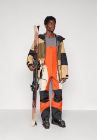 Suusariietus, mis sisaldab mitmevärvilist jakki, millel on beežid, rohelised ja mustad osad, ning erkoranže pritsmed. Reguleeritavad kinnitused ja tõmblukud on nähtavad.