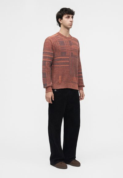 Jeune homme debout de profil, portant un pull orange à motifs, un pantalon large noir et des chaussures marron sur fond blanc.