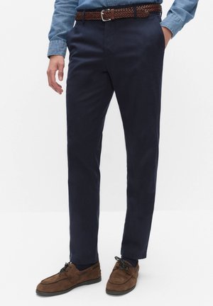 Homme portant un pantalon bleu foncé, une ceinture tressée marron, des chaussures en daim marron à lacets et une chemise en jean, une main dans la poche.