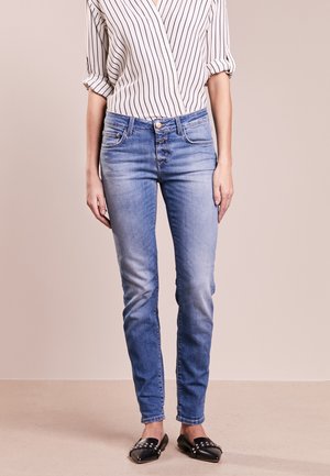 Vrouw die blauwe slim-fit jeans draagt, een witte blouse met zwarte verticale strepen, opgerolde mouwen, en zwarte loafers met studs, tegen een effen achtergrond.