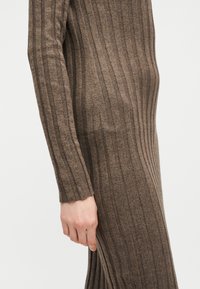 Gros plan d'une personne portant une robe en maille marron côtelée à manches longues et ajustée, la main détendue le long du corps.