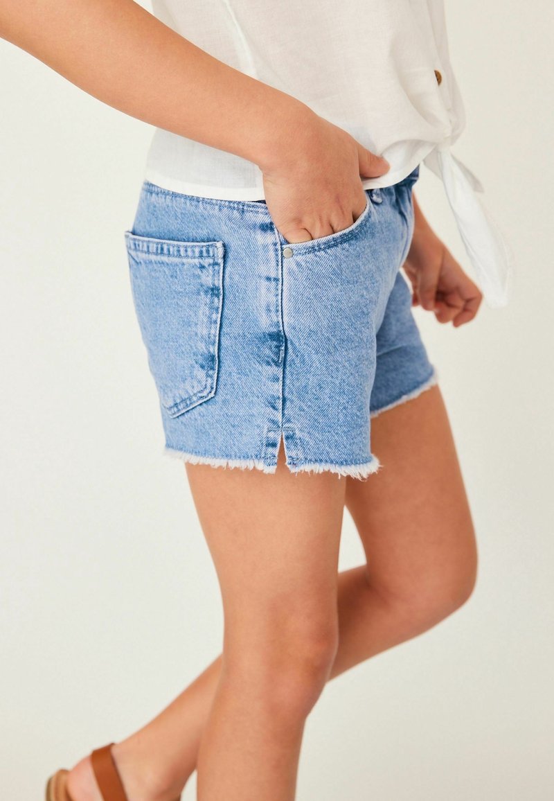 Personne portant un short en jean bleu clair avec des bords effilochés et une chemise blanche nouée, debout avec une main dans la poche.