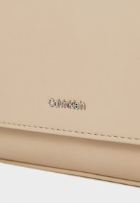 Portefeuille synthétique beige Calvin Klein avec une surface texturée et un logo argenté. Comprend des coutures visibles le long des bords.