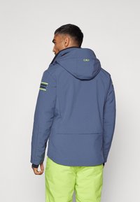 Chaqueta azul con capucha de textura suave, con acentos de rayas negras y verde neón en las mangas, combinada con pantalones verdes brillantes.