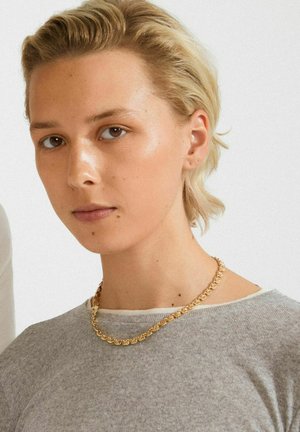Person mit kurzen blonden Haaren, der eine goldene Kettenhalskette und ein hellgraues geripptes Shirt trägt, leicht nach links geneigt vor einem schlichten Hintergrund.