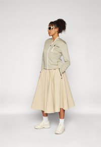 Lysebeige zip-up hettegenser med en liten brystlomme, kombinert med et flott kremfarget skjørt og hvite joggesko. Designet er avslappet og moderne.
